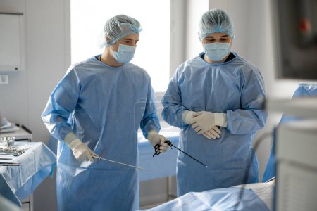 Téléchargez les photos : Deux chirurgiens en uniforme se tiennent debout avec des endscopes se préparant à une intervention mini-invasive pour un patient en salle d'opération. Concept de traitement endoscopique assisté par ordinateur - en image libre de droit