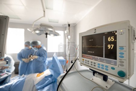 Photo for Surgery treatment in progress. Heart rate monitor on a foreground and surgeons working behind. Concept of monitoring the patients condition during surgery - Royalty Free Image