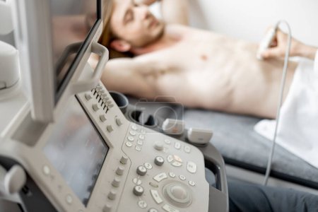 Téléchargez les photos : Jeune homme lors d'une échographie diagnostique du bas-ventre, gros plan sur la machine devant. Le concept de diagnostic par ultrasons et la santé des hommes - en image libre de droit