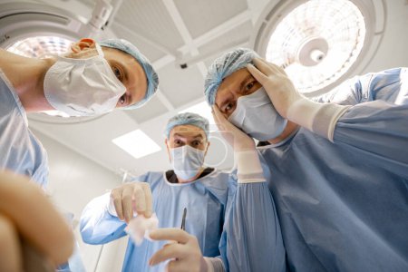 Foto de Los cirujanos asustadizos miran el lugar de la intervención quirúrgica con horror durante una operación, vista desde abajo. Descubrimiento de lo terrible durante la disección quirúrgica - Imagen libre de derechos