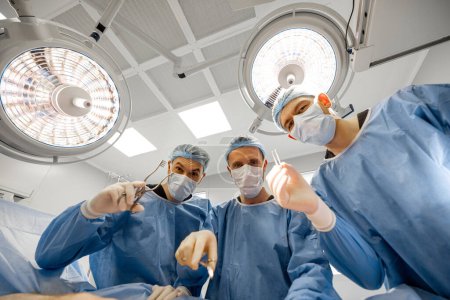 Téléchargez les photos : Portrait de trois chirurgiens effrayants avec des outils chirurgicaux regardez la caméra dans la salle d'opération - en image libre de droit