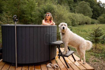 Téléchargez les photos : Femme se baignant dans un bain à remous extérieur tout en se reposant avec son chien mignon à la maison dans les montagnes. Concept de loisirs et de passer du temps libre avec des animaux sur la nature - en image libre de droit