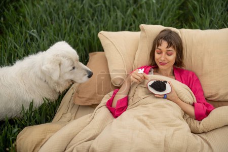 Foto de Mujer disfruta de dulce postre mientras se acuesta en la cama con un perro al aire libre. Mujer disfruta de la mañana en ropa de cama suave en un campo verde. Dulces sueños concepto - Imagen libre de derechos