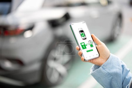 Photo for Woman holds a smartphone with running application for vehicle charging, electric car charging on background, close-up - Royalty Free Image