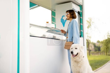 Foto de Mujer joven eligiendo helado para comprar en una hermosa tienda mientras pasea con su perro en un parque. Pasar tiempo de verano junto con mascotas al aire libre - Imagen libre de derechos