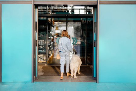 Foto de Mujer con perro blanco que entra en el supermercado con hermosas puertas correderas de color turquesa. Concepto de instituciones públicas o tiendas que admiten mascotas - Imagen libre de derechos