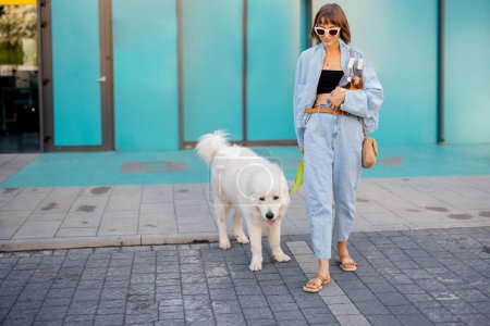 Téléchargez les photos : Femme allant avec son chien blanc mignon hors d'un supermarché, portant des bouteilles de vin devant les portes d'un magasin. Concept de firendship avec animaux domestiques et style de vie - en image libre de droit