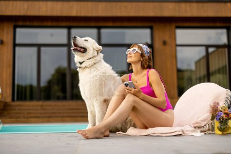 Téléchargez les photos : Femme étreint avec son chien blanc mignon tout en se relaxant près de la maison avec une piscine. Amitié avec les animaux domestiques et concept de vacances d'été - en image libre de droit