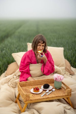 Foto de Mujer joven disfruta del desayuno de la mañana, comer pastel dulce mientras está acostado en la cama en el campo al aire libre. Concepto de dulces sueños y buenos días - Imagen libre de derechos