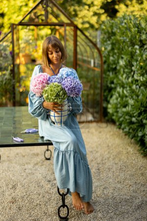 Téléchargez les photos : Portrait of a young woman in dress standing with beautiful bouquet in vase in garden. Floristry and gardening concept - en image libre de droit