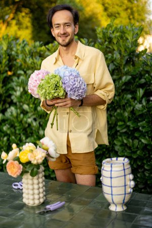 Téléchargez les photos : Portrait d'un homme joyeux arrange un bouquet avec des fleurs coupées dans le jardin. Homme fleuriste travaille dans son jardin à l'extérieur - en image libre de droit