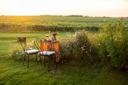 Foto de Beautiful green lawn with flowers and dining table for two during the sunset. Romantic picnic on nature - Imagen libre de derechos