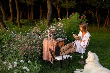 Foto de Mujer comiendo la cena mientras está sentada con su perro junto a la mesa en un césped de flores al aire libre - Imagen libre de derechos