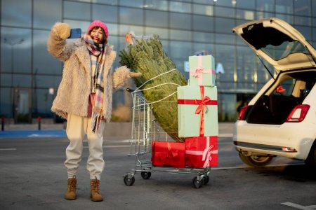 Foto de Joven mujer alegre hablando por teléfono mientras está de pie con carrito de compras lleno de regalo y árbol de Navidad cerca de coche en el estacionamiento de un centro comercial - Imagen libre de derechos