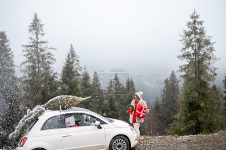 Téléchargez les photos : Jeune femme avec une boîte cadeau près de sa voiture avec arbre de Noël sur un toit sur neige, voyageant en véhicule haut dans les montagnes pendant les vacances d'hiver - en image libre de droit
