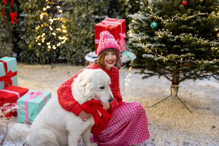 Téléchargez les photos : Portrait d'une femme avec arbre de Noël et étreintes boîte cadeau avec son adorable chien à la cour magnifiquement décorée sur la neige. Concept de vacances d'hiver heureuses et de magie - en image libre de droit