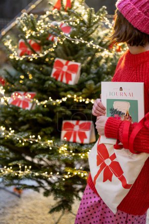 Téléchargez les photos : Femme près de l'arbre de Noël décoré de grands arcs rouges, tenant sac et magazine sur le thème du Nouvel An. Concept de décoration pour des vacances d'hiver - en image libre de droit