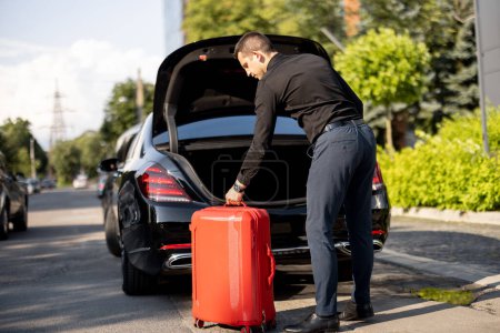 Photo for Chauffeur of a business-class car packs red suitcase into a trunk on the street near an office building. Concept of business trips, personal driver or luxury taxi - Royalty Free Image