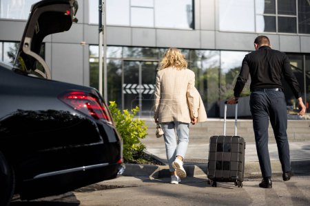 Téléchargez les photos : Chauffeur aide une femme d'affaires à transporter sa valise dans un immeuble, marchant ensemble, taxi de luxe en face. Concept de voyages d'affaires, idée d'un service de transfert de voiture de luxe - en image libre de droit