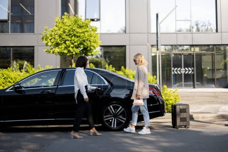 Photo for Female chauffeur opens a car door for a business lady going to sit, man packs a suitcase into a car trunk. Concept of luxury car transfer service, business trips and taxi - Royalty Free Image