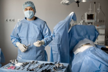 Téléchargez les photos : Portrait d'un chirurgien en uniforme avec des instruments médicaux stériles prêts pour la chirurgie en salle d'opération. Concept de traitement invasif - en image libre de droit