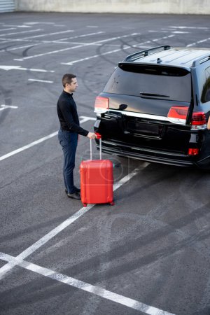 Photo for Male chauffeur standing with a suitcase near car on parking lot - Royalty Free Image