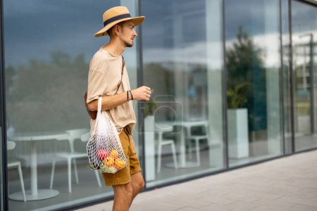 Téléchargez les photos : Élégant homme au chapeau marche avec un sac en filet plein de légumes frais près du supermarché à l'extérieur. Concept de style de vie durable - en image libre de droit