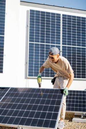 Téléchargez les photos : Homme installant des panneaux solaires sur le toit de sa maison, cellule à visser sur le toit plat pour l'autoconsommation. Concept d'énergies renouvelables et de durabilité - en image libre de droit
