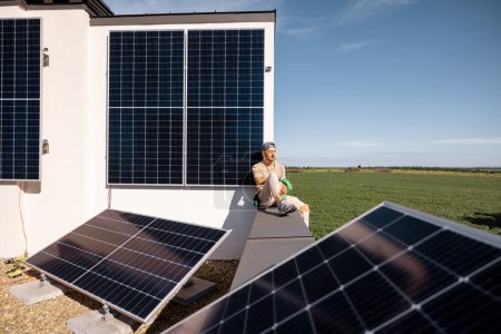 Foto de Hombre sentado en una azotea de su casa con una central solar instalada en ella. Energía verde renovable y concepto de sostenibilidad - Imagen libre de derechos