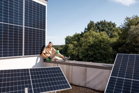 Foto de Hombre sentado en una azotea de su casa con una central solar instalada en ella. Energía verde renovable y concepto de sostenibilidad - Imagen libre de derechos