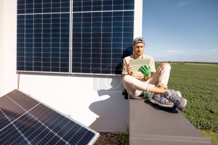 Téléchargez les photos : Homme assis sur un toit et utilisant une tablette numérique de surveillance de la production de la centrale solaire installée sur sa propriété. Concept de technologies modernes et mode de vie durable - en image libre de droit