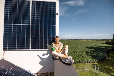Téléchargez les photos : Homme assis sur un toit et utilisant une tablette numérique de surveillance de la production de la centrale solaire installée sur sa propriété. Concept de technologies modernes et mode de vie durable - en image libre de droit