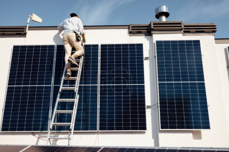 Foto de Hombre instalando paneles solares, subiendo en una escalera en el techo de su casa. Amplia vista angular en un tejado con central solar. Concepto de energía renovable y sostenibilidad - Imagen libre de derechos