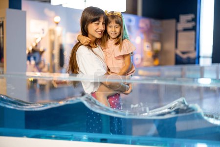 Foto de Mamá con hija pequeña aprende física interactivamente en un modelo que muestra fenómenos físicos mientras visita un museo de ciencias. Concepto de entretenimiento y aprendizaje infantil - Imagen libre de derechos