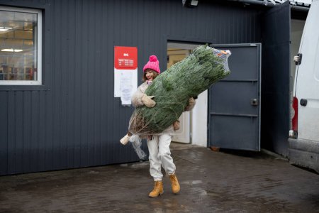 Foto de Mujer joven lleva el árbol de Navidad envuelto mientras sale de la oficina de correos entregado desde la tienda en línea. Concepto de entrega y compras en línea en vacaciones de invierno - Imagen libre de derechos