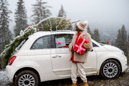 Téléchargez les photos : Jeune femme se tient avec une boîte cadeau près de sa voiture avec arbre de Noël sur un toit, voyageant en voiture sur les montagnes pendant les vacances d'hiver - en image libre de droit
