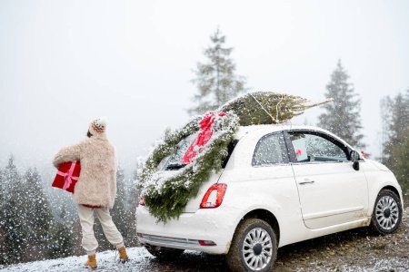 Photo for Young woman enjoys great view on pine forest on a snowfall, while traveling by car highly in the mountains during winter holidays. Vehicle with Christmas tree on rooftop and wreath behind - Royalty Free Image