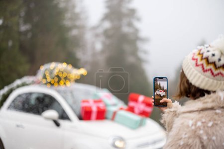 Foto de Mujer hace una foto en el teléfono de hermoso coche decorado con regalos y árbol de Navidad en la azotea, mientras viaja en vehículo en las montañas en las vacaciones de invierno - Imagen libre de derechos