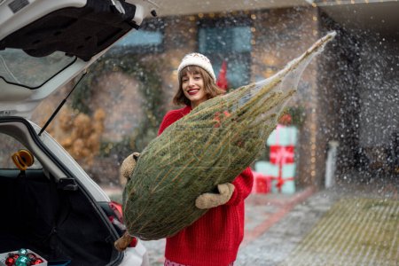 Foto de Young cheerful woman in red sweater and hat carries wrapped Christmas tree near car at porch of her house decorated for winter holidays. Concept of preparing and decorations for New Years holidays - Imagen libre de derechos