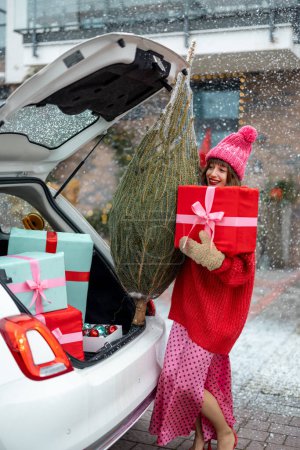 Téléchargez les photos : Young cheerful woman in red sweater and hat with wrapped Christmas tree and gift box near car at porch of her house decorated for winter holidays. Concept of preparing for a New Year - en image libre de droit
