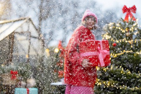 Photo for Portrait of a young cute woman in red sweater and hat stands with a gift box near Christmas tree at beautifully decorated backyard during a snow fall. Concept of Winter magic and holidays - Royalty Free Image