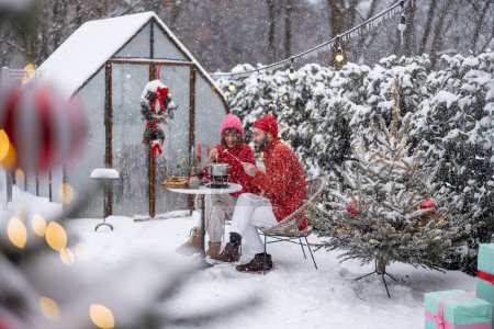 Man and woman have romantic dinner with fondue, while sitting together by the table at beautifully decorated snowy backyard. Young family celebrating winter holidays outdoors