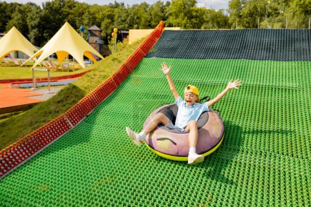 Téléchargez les photos : Garçon heureux descend dans le tube gonflable de traîneau sur une piste d'été, s'amusant au parc d'attractions. Concept de vacances d'été et de divertissement pour enfants - en image libre de droit
