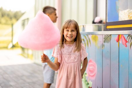 Foto de Niña feliz sostiene caramelo de algodón rosa mientras visita el parque de atracciones durante unas vacaciones de verano - Imagen libre de derechos