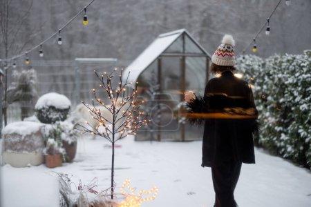 Téléchargez les photos : Femme en vêtements de nuit et chapeau sort sur son jardin enneigé le matin pendant les vacances d'hiver - en image libre de droit