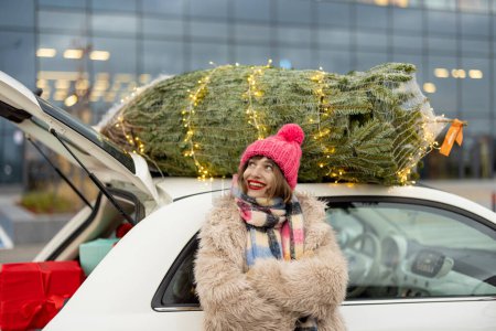 Foto de Mujer alegre soñando para unas felices vacaciones, mientras que de pie cerca del coche con el árbol de Navidad por encima. Chica de compras para unas vacaciones de Año Nuevo en el centro comercial - Imagen libre de derechos