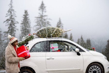 Téléchargez les photos : Jeune femme se tient avec une boîte cadeau près de sa voiture avec arbre de Noël sur un toit, voyageant en voiture sur les montagnes pendant les vacances d'hiver - en image libre de droit