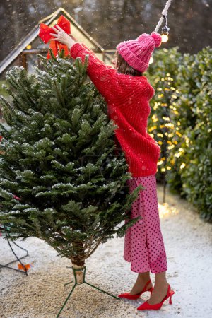 Téléchargez les photos : Jeune femme en rouge décore arbre de Noël luxuriant avec des arcs festifs à l'arrière de sa maison sur la chute de neige, se préparant pour des vacances d'hiver - en image libre de droit