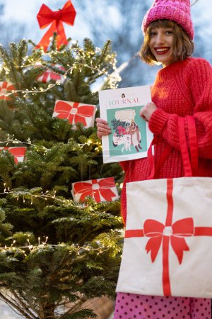 Téléchargez les photos : Femme près de l'arbre de Noël décoré de grands arcs rouges, tenant sac et magazine sur le thème du Nouvel An. Concept de décoration pour des vacances d'hiver - en image libre de droit
