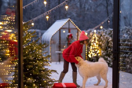 Téléchargez les photos : Woman plays with her cute white dog at beautifully decorated and snowy yard, celebrating and having fun together during winter holidays. Concept of happy winter holidays, magic and love - en image libre de droit